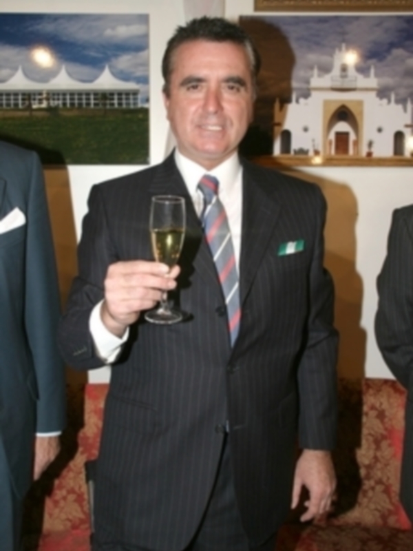 Un hombre en traje negro con una corbata celeste y un pequeño emblema verde sostiene un vaso de champán. Fondo con dos cuadros, uno con una imagen de edificios y el otro con un palacio. Suelo rojo y alfombra en la parte inferior.