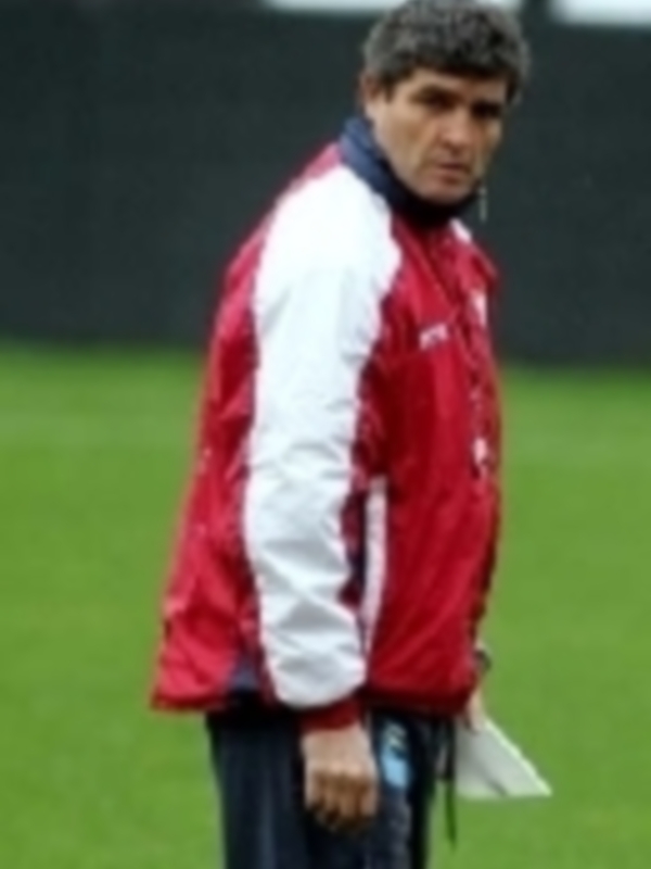 Un hombre con una chaqueta azul y roja, posiblemente un entrenador o técnico de fútbol, observa algo en el campo.