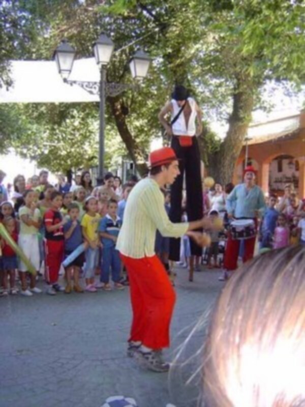 Una escena callejera con un payaso de alto en pantalones rojos y una camisa amarilla, interactuando con un niño pequeño en el centro. Al fondo, se observa a otros niños y adultos disfrutando de la actuación en un ambiente festivo.