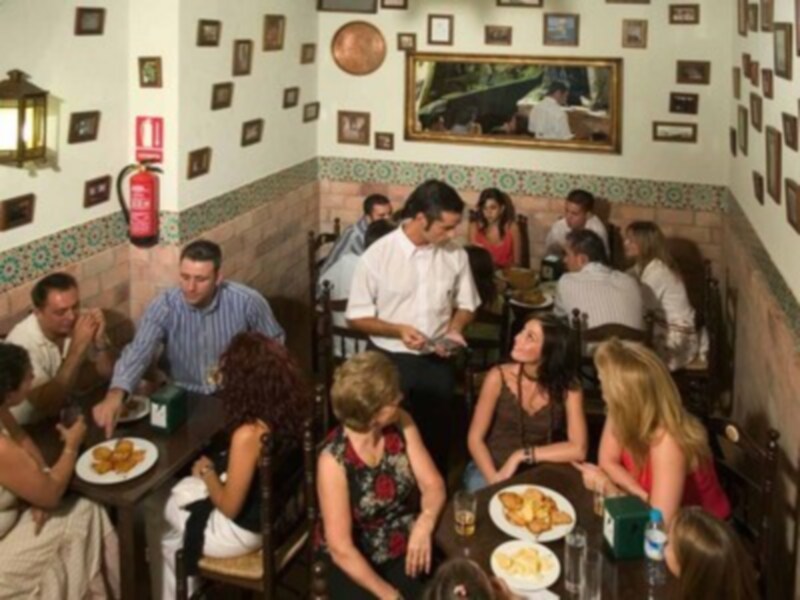 Una escena en un restaurante con mesas llenas de comensales. En la pared se pueden ver fotos y una esfera. La decoración incluye azulejos blancos y negros.