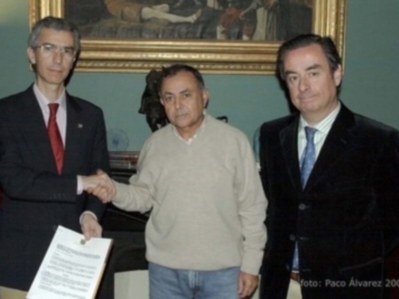 Tres hombres en trajes, uno con corbata roja y otro con corbata azul, estrechan la mano mientras el tercero sostiene un documento. En el fondo, una pintura de dos figuras se adorna la pared.
