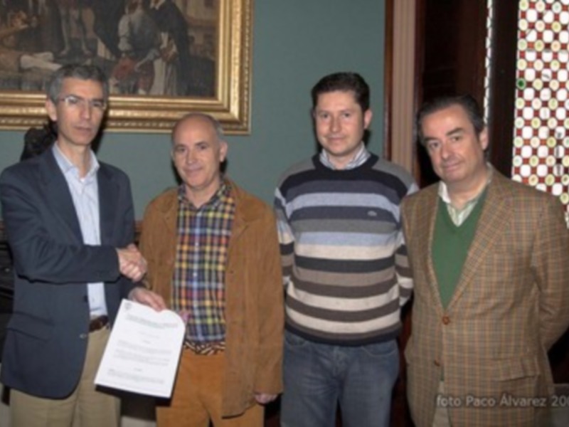 Un grupo de personas posando para una foto, dos hombres están sosteniendo un documento mientras otros dos observan. La imagen está firmada por Paco Álvarez 2007.