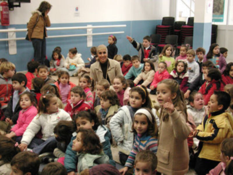 Niños y niñas en una escuela, algunos con abrigos y gorros. Una maestra adulta está entre ellos, posiblemente enseñando o interactuando con los niños.