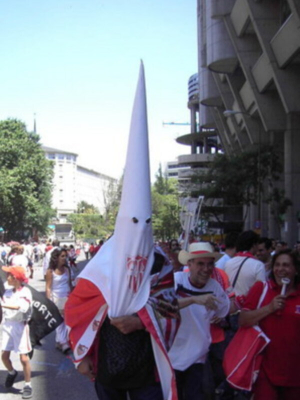 Una multitud de personas camina por una calle con edificios modernos al fondo. Algunos llevan banderas blancas con rojo y negro, mientras otros sostienen pancartas. La escena parece ser una manifestación o protesta en un entorno urbano.