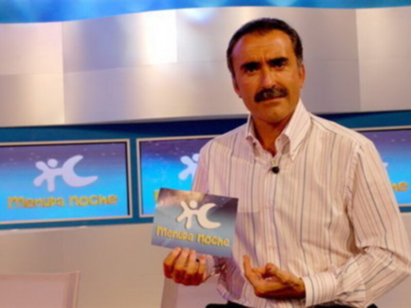 Un hombre con barba y bigote sostiene un tarjetón que dice "Mancha noche". El fondo muestra pantallas de televisión con la misma imagen.