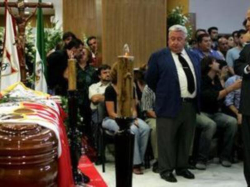 Una caja funeraria con banderas ondeando, rodeada de personas en trajes formales. En el fondo, una persona con un sombrero y otro con una bufanda negra. La escena parece ser un funeral o ceremonia funeraria en un entorno interior con decoraciones y velas.