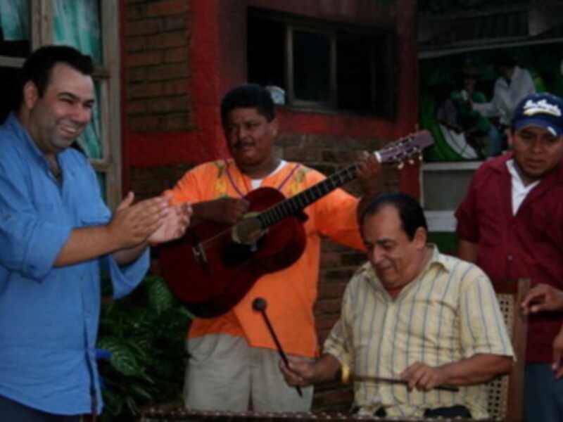 Un grupo de personas disfruta de una reunión musical, con un hombre en el centro tocando la guitarra mientras otros aplauden y observan. La escena parece ser en un ambiente informal, posiblemente una fiesta o evento local.