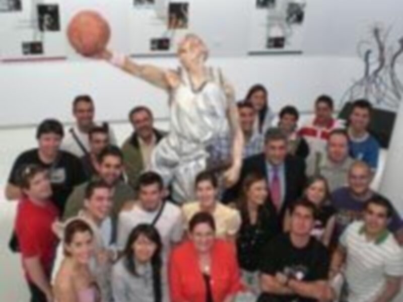 Una escultura de un jugador de baloncesto en acción, con una pelota en la mano, rodeada por una multitud de personas sonrientes. La escultura y las personas están en un entorno interior con paredes blancas y una iluminación suave.