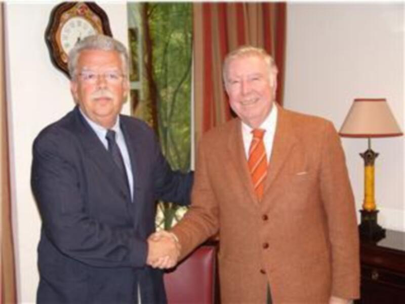 Dos hombres en trajes, uno con corbata azul y el otro con una corbata naranja, están estrechando la mano en un ambiente formal. La imagen muestra una sala con cortinas rojas y un reloj de pared en el fondo.
