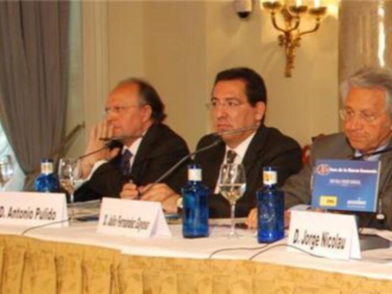 Tres hombres sentados en una mesa de conferencia con nombres escritos, sosteniendo copas y botellas de agua.
