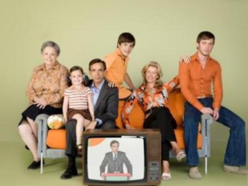 Una familia de cinco personas, compuesta por una abuela, un padre y tres hijos, posan frente a un televisor antiguo. La abuela y el padre están sentados en una silla, mientras los hijos se sientan en otra. Todos lucen atuendos de colores brillantes, y la abuela tiene un sombrero. La escena se desarrolla en un fondo verde claro, con una silla de color naranja.
