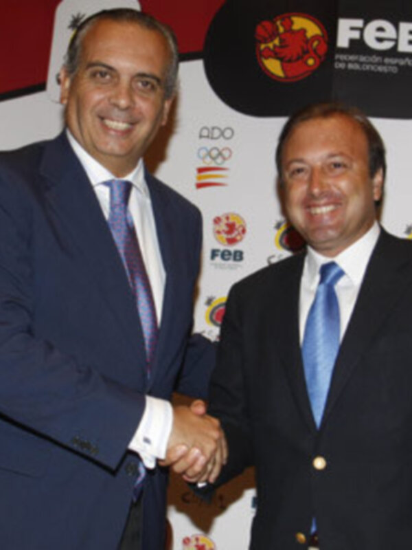 Dos hombres en trajes se saludan en un evento deportivo, con logos de la Federación Española de Baloncesto (FEB) y otras organizaciones en el fondo.