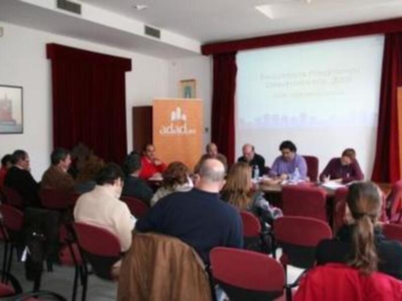 Presentación en sala de conferencias con asistentes sentados, una pantalla proyectando un documento y la marca ADAC en el fondo.