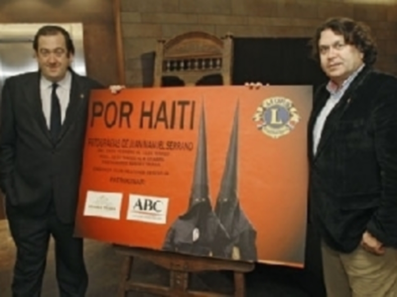 Dos hombres posan junto a un cartel que dice 'POR HAITI' y muestra una imagen de una catedral, apoyando una causa benéfica.