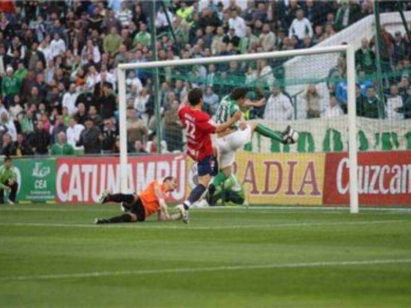 Un jugador de fútbol en acción, con el balón cerca del arco contrario. El portero está tendido en el suelo, mientras un jugador ataca con la pelota. El césped es verde y el fondo muestra a los espectadores en una grada.