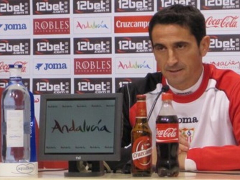 El jugador está en una conferencia de prensa con un fondo de anuncios de Coca-Cola y Joma. En la mesa hay una botella de agua, una botella de cerveza y un monitor con el texto "Andalucía".