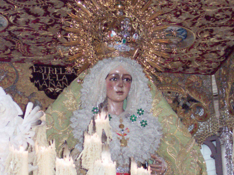 Una figura religiosa vestida con vestimentas de color verde y dorado, rodeada por adornos elaborados. La figura lleva un tocado con flores y una corona dorada, y está rodeada por detalles artísticos y texturizados en el fondo.