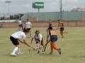 Jugadoras de hockey en acción durante un partido, con una señal de publicidad visible en el fondo.