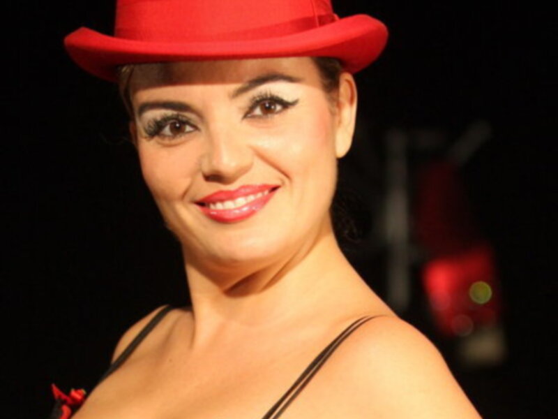 Una mujer con un sombrero rojo y una vestimenta de estilo burlesco, posando en un escenario iluminado.