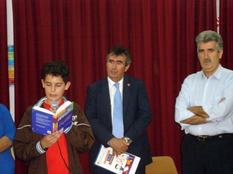 Un niño sostiene un libro mientras dos hombres observan detrás de una cortina roja.