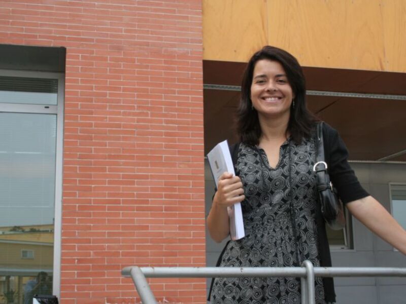 Una mujer sonríe mientras sostiene un libro en un entorno urbano con edificios de ladrillo y madera.
