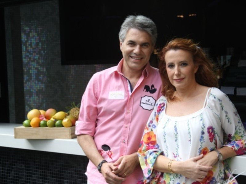Un hombre y una mujer sonríen en frente de una barra de frutas. El hombre lleva un camisa rosa y la mujer una blusa con flores multicolores. La barra de frutas incluye naranjas, limones y una piña. El fondo muestra un mosaico de colores brillantes.