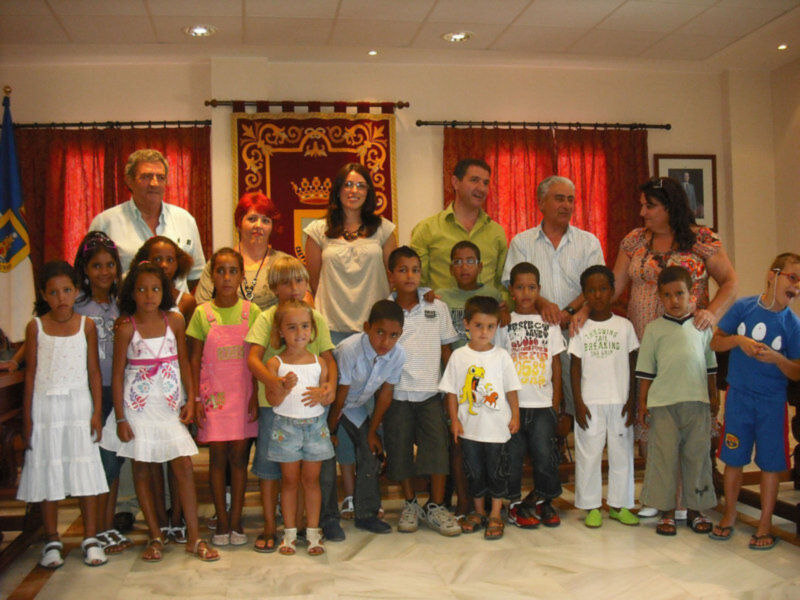 Un grupo de niños y adultos posan en una sala con cortinas rojas, un escudo y una bandera. La imagen muestra a niños de diferentes edades y estilos vestimentarios, posando juntos en una escena formal.