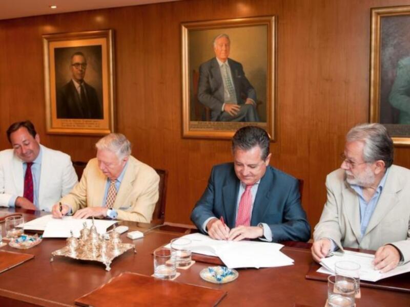 Cuatro hombres en trajes están sentados a una mesa, firmando documentos. En la pared detrás de ellos hay retratos de hombres. La imagen parece ser de una reunión formal o negociación.