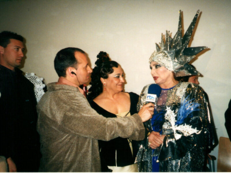 Una persona vestida con un traje brillante y una corona de plumas interactúa con otros dos individuos, uno de ellos sosteniendo un micrófono. La escena parece ser parte de una celebración o evento formal, con vestimentas elegantes y un ambiente festivo.
