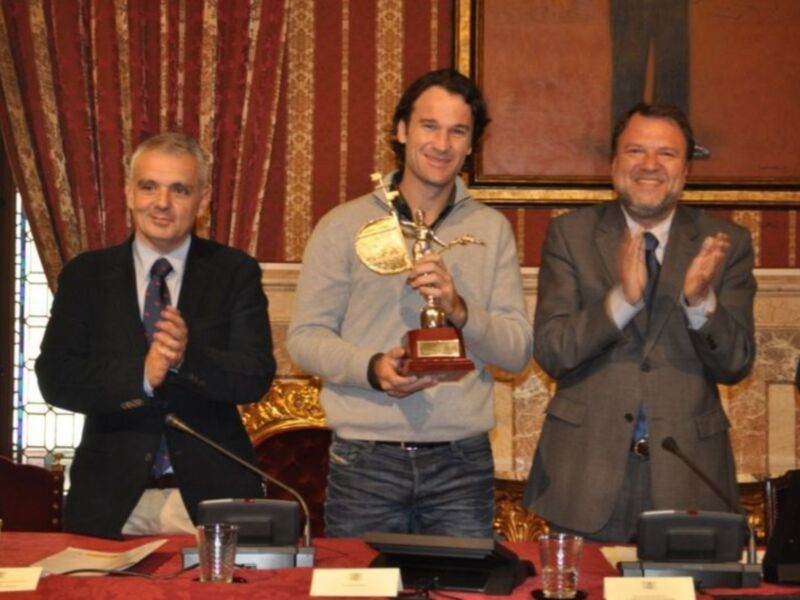 Tres hombres celebran en una sala de conferencias, uno sostiene un trofeo mientras los otros aplauden. La decoración y el ambiente sugieren una ceremonia formal o de reconocimiento.