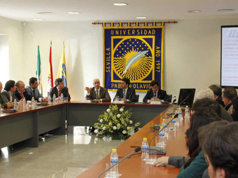 Sala de conferencias con una bandera universitaria en el fondo. Miembros del consejo directivo se sientan alrededor de una mesa con banderas nacionales y un escudo en el centro. La pantalla a la derecha muestra información técnica.