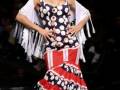 Una modelo muestra un vestido flamenca con estampado de lunares rojos y blancos, complementado por una falda roja con franjas negras. La pieza incluye un escote en V y mangas de encaje, con una falda que se despliega al moverse. La pieza es complementada por un tocado tradicional con una flor roja y un escote en V.