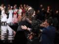 Una mujer en un vestido negro con flores abraza a una otra persona, ambos están en el centro de la imagen. La escena parece ser un evento formal o una pasarela, con luces y público en el fondo.