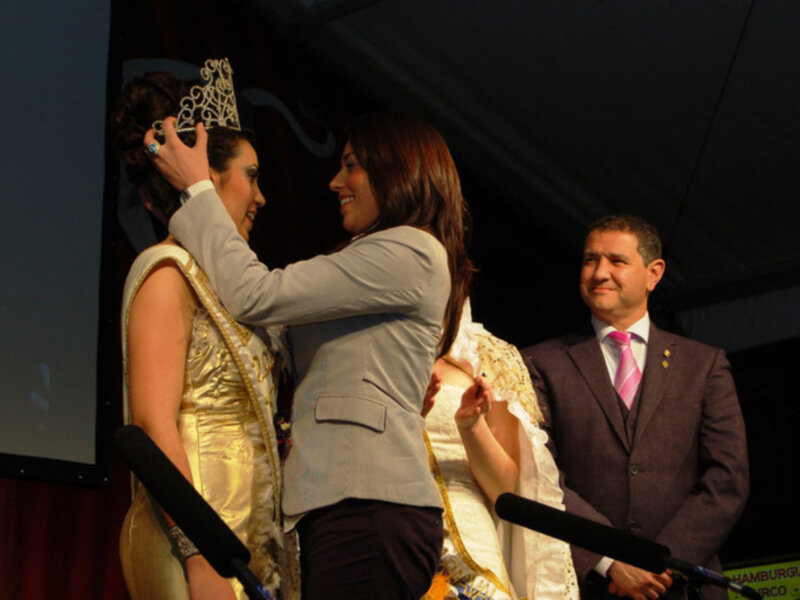 Una mujer en un vestido dorado y corona de diamantes se encuentra en el escenario, mientras otra mujer le coloca la corona. Un hombre en traje y corbata se encuentra detrás de ellas, sonriendo.
