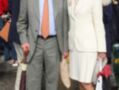 Una pareja formal posa juntos, el hombre en traje gris y corbata naranja, la mujer en abrigo blanco. Detalle de la imagen: hombre con sombrero y paraguas, mujer con zapatos rojos.
