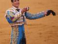 Un torero en traje tradicional de color azul y dorado, con un fondo de arena rojiza.