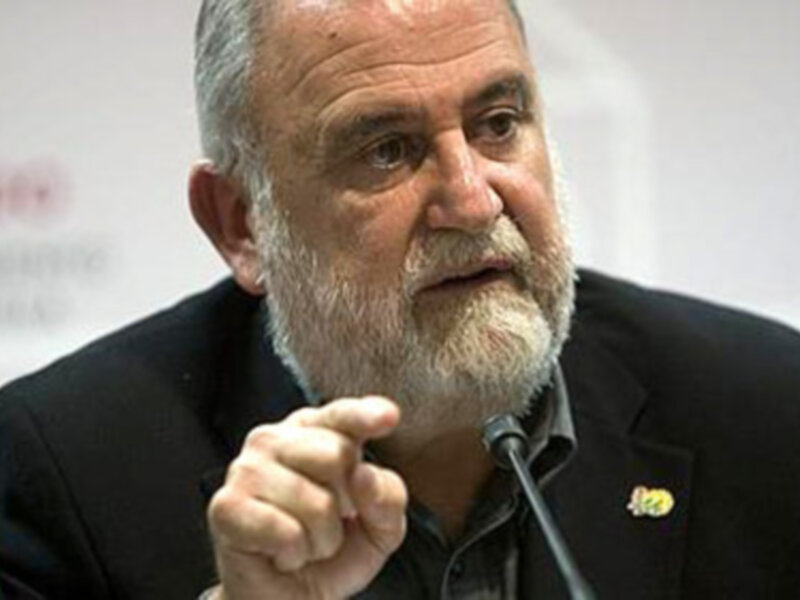 Un hombre con barba blanca y cabello gris, vestido de negro, hablando en un evento formal.