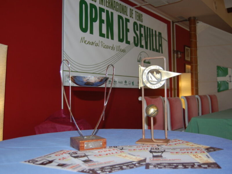 Tres trofeos de tenis en una mesa con un cartel que dice "Open de Sevilla" y un banner de la Federación Internacional de Tenis.