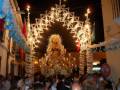 Fiesta religiosa nocturna con una imagen de la Virgen en el centro, rodeada por luces y adornos. Banderas y decoraciones festivas iluminan el ambiente.