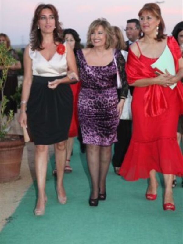 Una mujer con vestido negro y blusa blanca, otra en un vestido morado con estampado de leopardo y una tercera en un vestido rojo largo. Todos caminan sobre un camino verde, con una pila de flores en el fondo y una persona detrás.