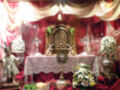 Altar con adornos florales y figuras religiosas, decorado con telas rojas y doradas.