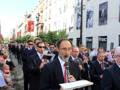 Una procesión religiosa en una calle con edificios blancos y rojos. Un hombre toca un instrumento musical mientras otros participan en el evento.
