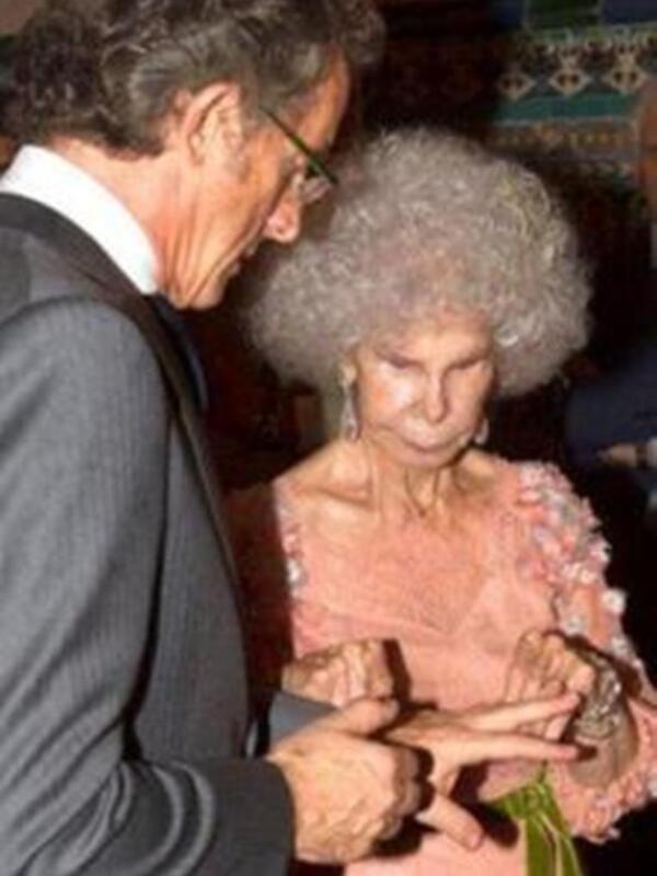 Una anciana con cabello blanco y una blusa rosa, mirando hacia abajo mientras un hombre en traje le muestra algo en la mano.