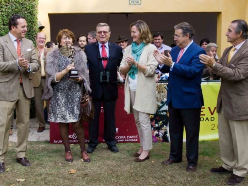 Una celebración formal con personas vestidas de manera elegante, posiblemente en un evento deportivo o empresarial. La imagen muestra a varias personas, incluyendo una mujer con un vestido de lentejuelas y otro con un abrigo beige, posiblemente en una ceremonia o evento.