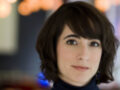 Una mujer con cabello corto y oscuro, vistiendo una camiseta de cuello alto azul. El fondo es borroso con luces brillantes que sugieren un ambiente moderno y acogedor.