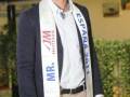 Un hombre sonríe mientras posa con un gran corbata blanca que dice "Mr. España 2011". Está vestido con una chaqueta azul y pantalones beige. La imagen está tomada en un área con vegetación y una estructura metálica al fondo.