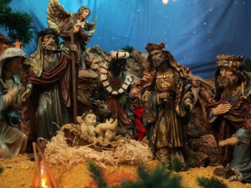 Escena navideña con figuras de madera representando a los Reyes Magos, el Niño Jesús en el nacimiento y otros personajes. La escena está iluminada con un fondo azul y hay detalles de paja y plantas verdes.