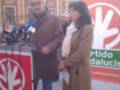 Dos personas, una hombre y una mujer, están en un evento público frente a un edificio histórico. El hombre está hablando mientras sostiene micrófonos y la mujer escucha atenta. Detrás de ellos hay un cartel con el logo del Partido Andalucista y una bandera roja.