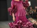 Una mujer en un desfile de moda flamenca, con un vestido fucsia de tul y volantes, complementado con pendientes grandes y una flor en el cabello.