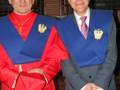 Dos hombres vestidos formalmente, uno con un traje rojo y otro con un chaleco azul. Ambos llevan insignias de honor en sus pechos y parecen estar en una ceremonia o evento importante.